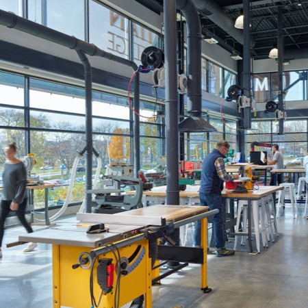 Modern fabrication lab with industrial woodworking equipment, large windows and students working at tables in a bright makerspace.